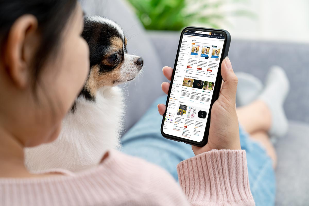 Composite photo of woman reclined on couch holding small dog and phone while looking up GPS dog tracker results on the Amazon website