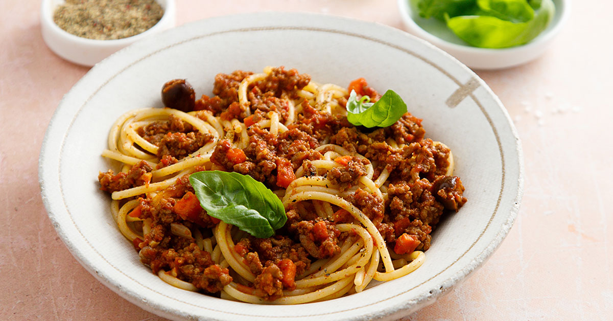 Photograph of a rustic white bowl of savory spaghetti topped with fresh basil leaves.