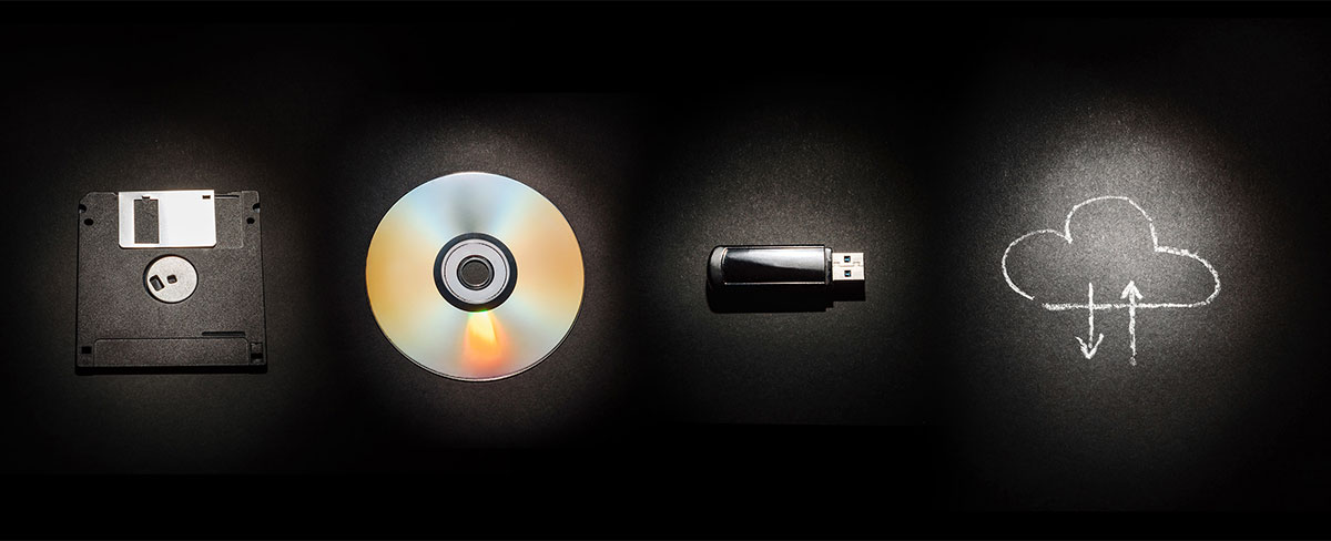 Floppy disc, CD, flash drive, and a chalk drawing of storing things in the cloud lined up on a black background.